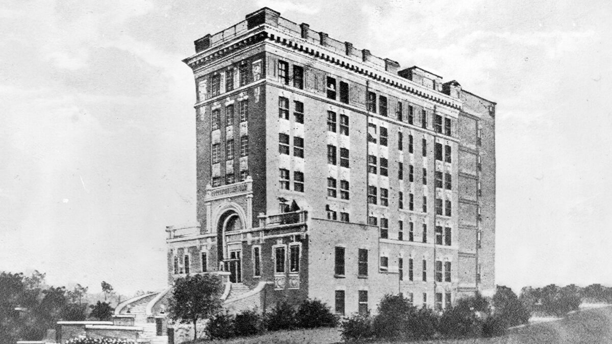 Historic multi-story brick building with arched entrance and rows of windows, surrounded by trees.